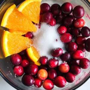 cranberries, orange wedges, and sugar in a food processor on a white countertop.